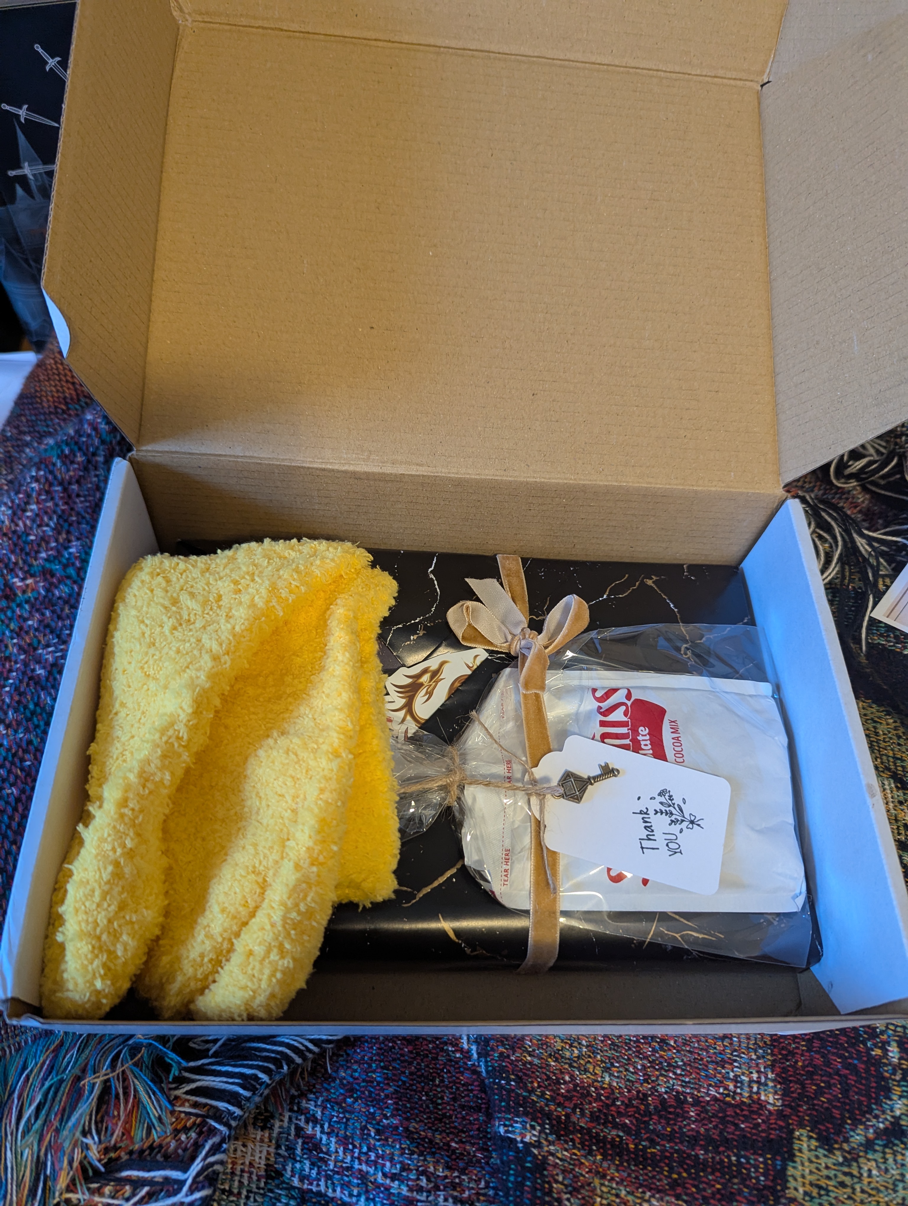 Image shows a box with a pair of fuzzy yellow socks, and a book wrapped up in wrapping paper with hot chocolate and a thank you tag.